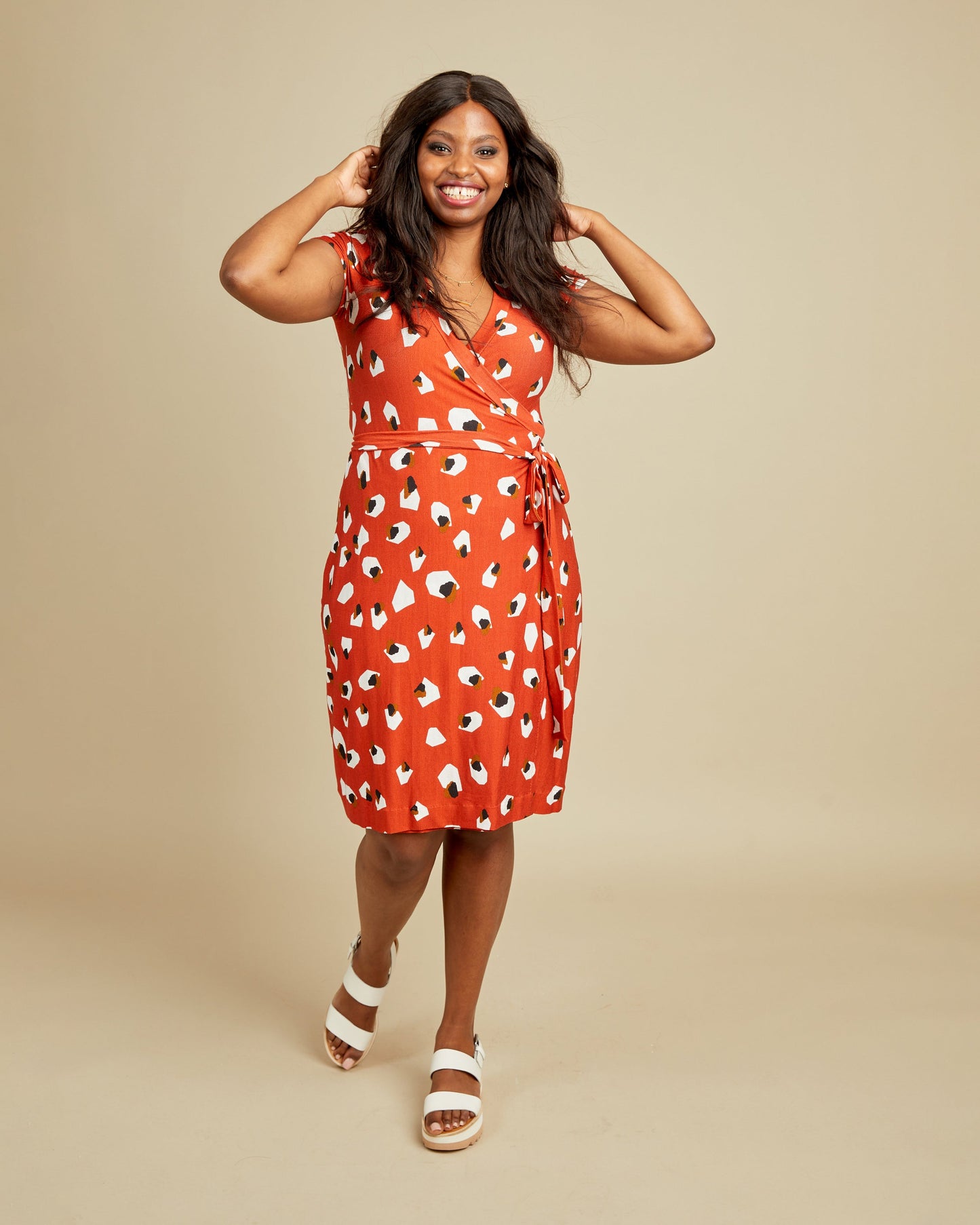 Woman wearing a red dress with a pattern on a beige background. This dress is the Appleton Wrap Dress sewing pattern from Cashmerette.