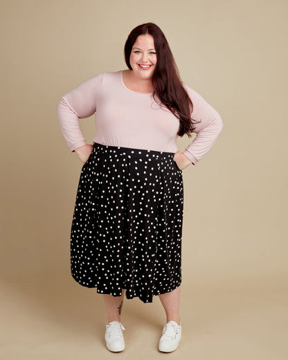 Woman wearing a pink top and black polka dot skirt on a beige background. This t-shirt is the Concord T-Shirt sewing pattern from Cashmerette.