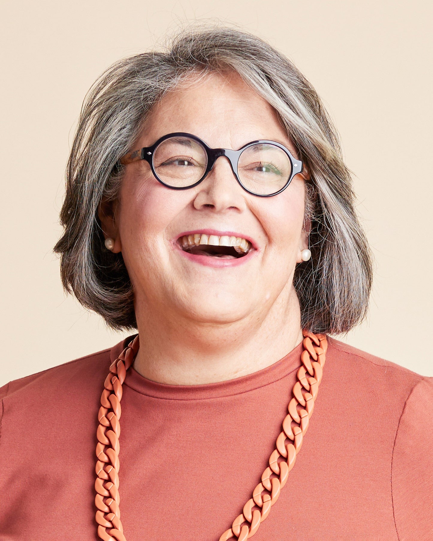 Person with gray hair wearing glasses and a brown shirt with a large orange necklace on a beige background. This is the Grafton Dress, Top & Skirt sewing pattern from Cashmerette.