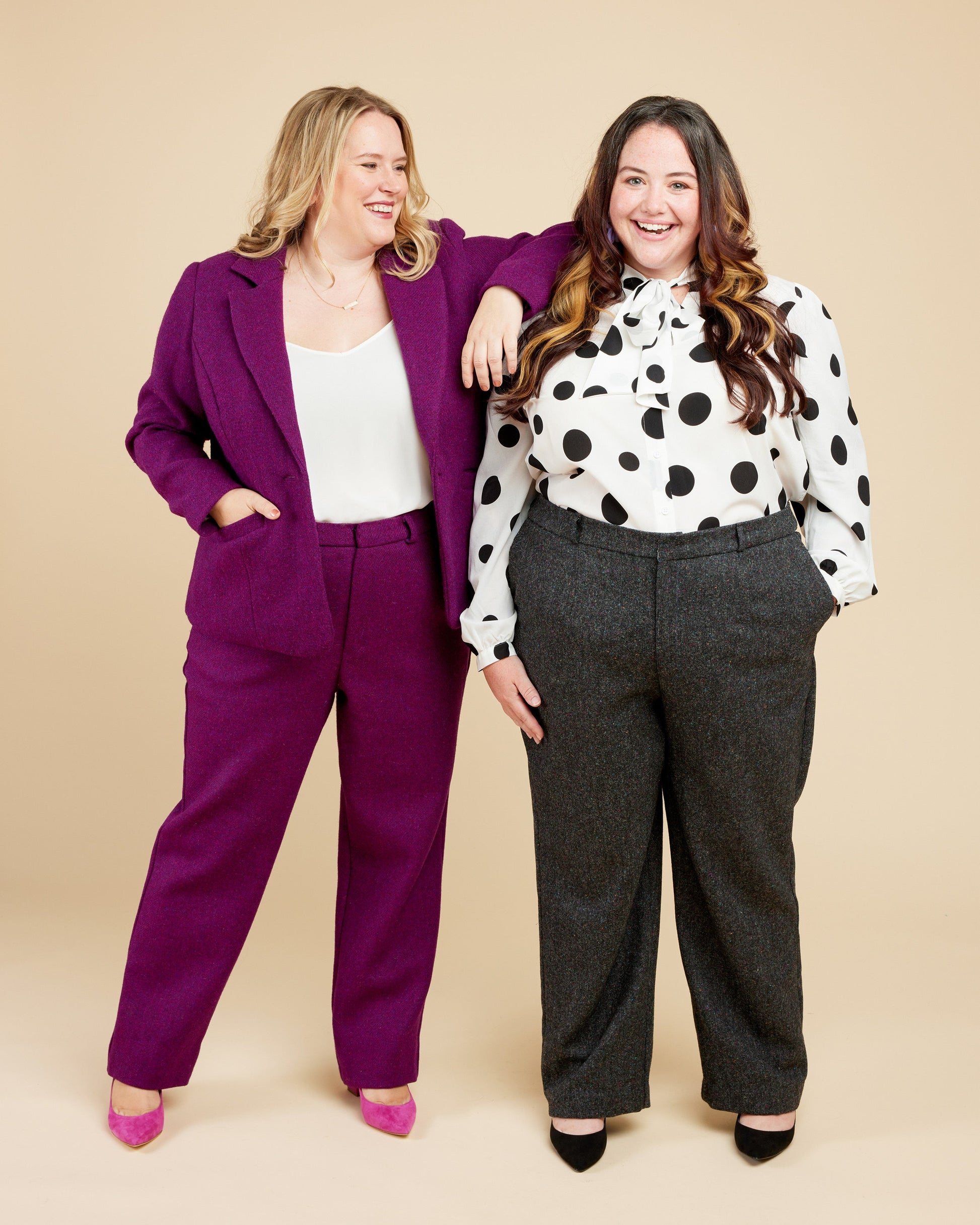 Two women posing together against a beige background, one in a purple suit and the other in a polka dot blouse and gray pants.. These pants are the Meriam Trousers sewing pattern from Cashmerette.