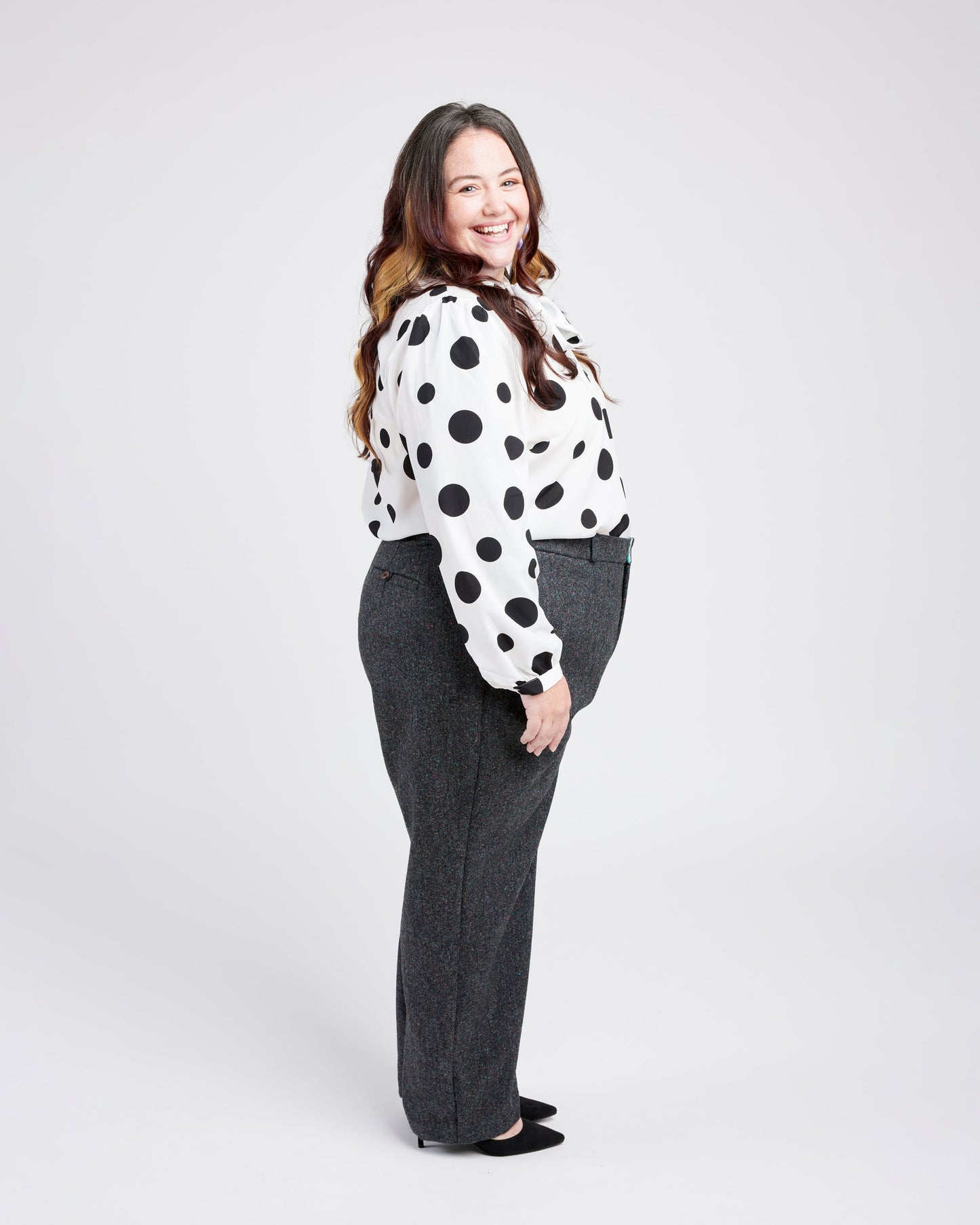 Woman wearing a polka dot blouse and gray pants on a white background. These pants are the Meriam Trousers sewing pattern from Cashmerette.