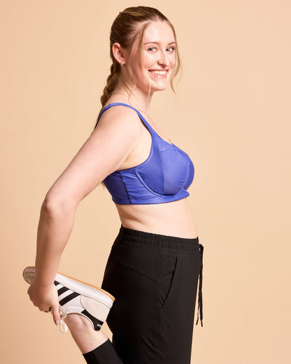 Woman wearing a blue sports bra and black leggings, holding white sneakers against a beige background. This is the Cashmerette Somerville Sports Bra sewing pattern for big boobs.