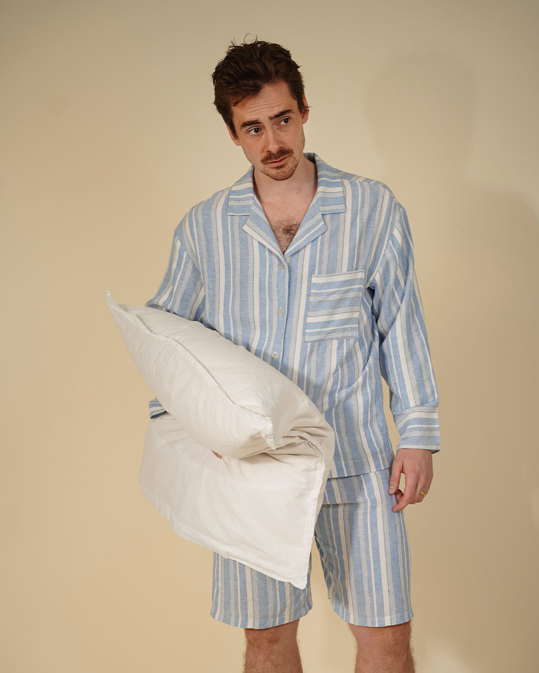 A man wearing a light blue and white vertically striped pajama set holds a white pillow against a solid beige studio background.
