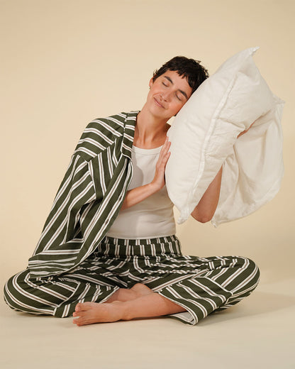 A woman sits cross-legged on a beige studio floor wearing a matching green and white striped pajama set, with a white pillow against her head.