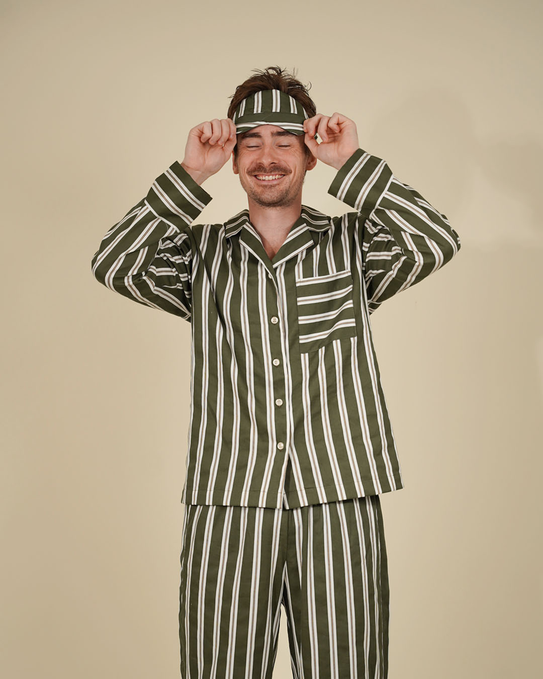 A man wearing a matching green and white vertically striped pajama set with a headband, posing with his hands on the headband against a solid beige background.