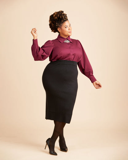 Woman wearing a burgundy blouse and black skirt on a beige background. This top is the Vernon Shirt sewing pattern from Cashmerette.