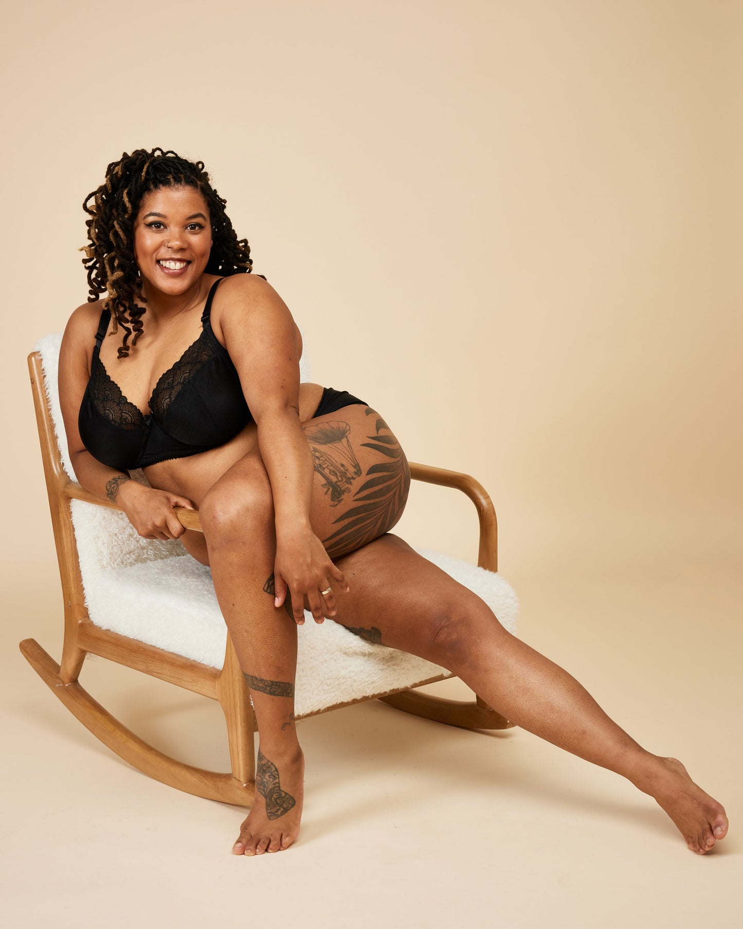 Woman sitting on a rocking chair wearing black lingerie against a beige background. This bra is the Willowdale Bra sewing pattern from Cashmerette.